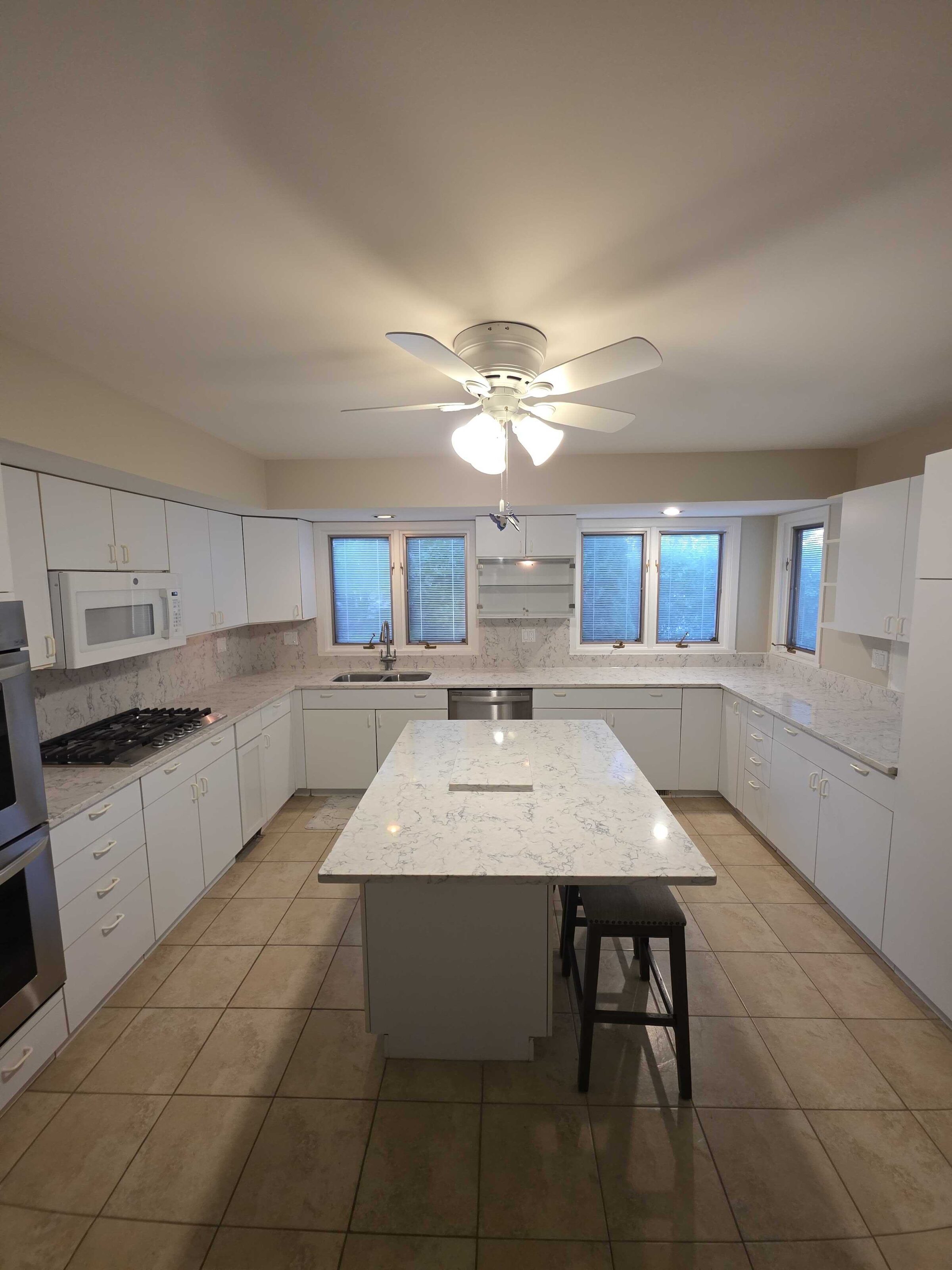 White kitchen with a marble-look quartz island, wall ovens to the left and windows along the back